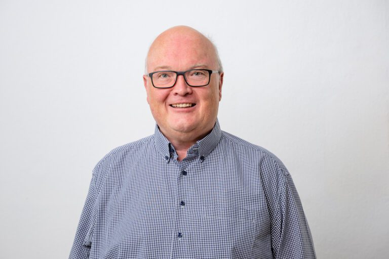 Ortschronist Claus Strobl. Porträt eines lächelnden Mannes mit Brille und kariertem Hemd vor weißem Hintergrund.
