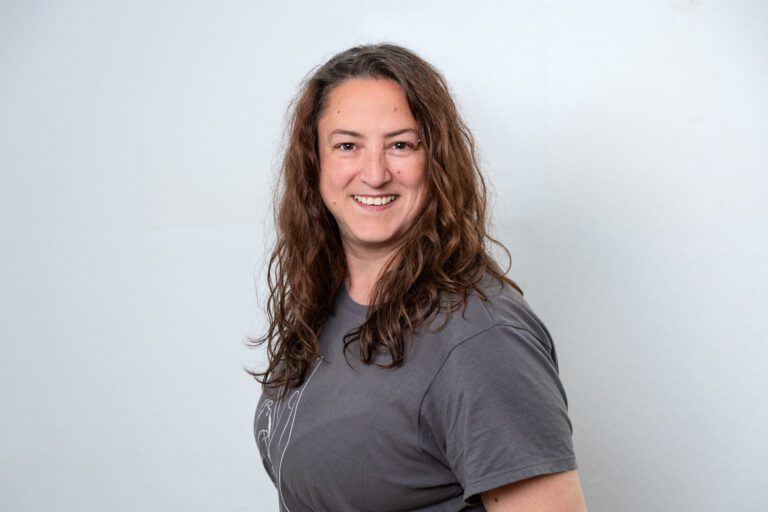 Erst, Melanie. Porträt einer Frau mit langen lockigen braunen Haaren im grauen Shirt.
